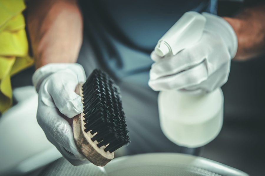 Nahaufnahme: behandschuhte Hände mit Handbürste und Reinigungsmittel über einem Waschbecken.