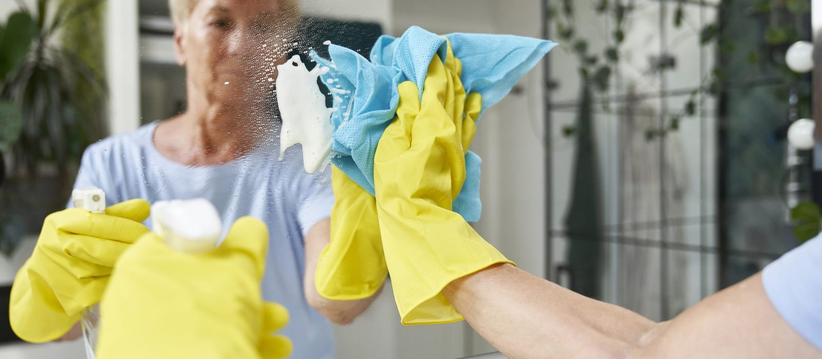 Eine Mitarbeiterin mit gelben Handschuhen reinigt eine eingeschäumte Spiegelfläche.
