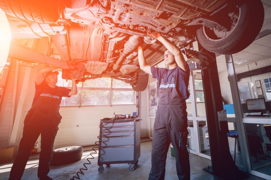 Zwei Mechaniker arbeiten von unten am Fahrwerk eines angehobenen Fahrzeugs, von links fällt warmes Licht in die Halle.